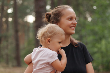 Side view portrait of smiling young adult Caucasian woman wearing black casual T-shirt holding baby daughter while enjoying beautiful nature in summer pine trees forest