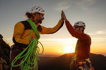 Two men high on a mountain, one of them is wearing a yellow jacket. They are high up on a mountain and are high fiveing each other - obrazy, fototapety, plakaty