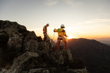 Two people are standing on a mountain, one of them is wearing a yellow jacket. The sun is setting in the background, creating a beautiful and serene atmosphere - obrazy, fototapety, plakaty