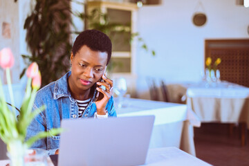 business black woman use cell smart phone while sitting at bistro remote working coding with laptop