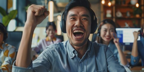 An office-based young Asian man contact center telemarketing agent cheers and punches the air. Excited businessman celebrating sales and meeting goals to win - obrazy, fototapety, plakaty