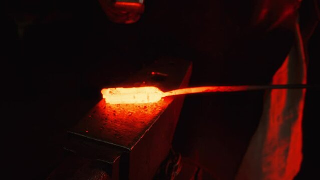 A blacksmith uses a hammer to shape a piece of red-hot metal in a forge on an anvil, creating a future product, sparks fly from blows. The process takes place in the darkness of the forge.