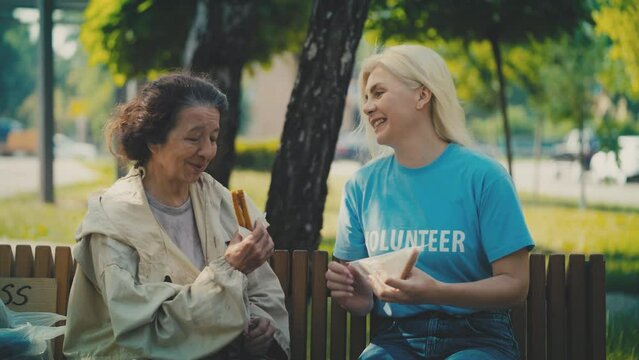 Volunteer giving sandwich to homeless senior woman in park, food donation