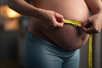 A close-up shot of an expectant pregnant woman measuring her round belly with a measuring tape