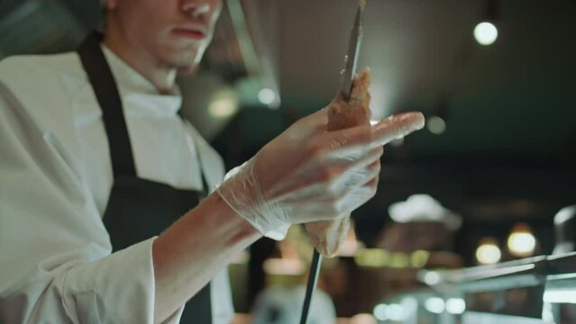 Young male chef in apron and gloves sticking ground meat to screwer while cooking kebab in restaurant kitchenYoung male chef in apron and gloves sticking ground meat to screwer while cooking kebab in 