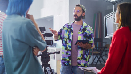 Young Caucasian Male Director With Beard And Glasses, Instructing Diverse Team On Film Set, Holding...