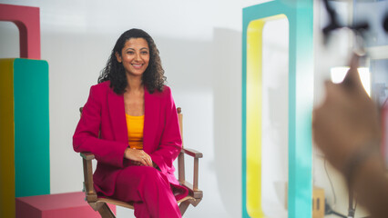 Young Middle Eastern Female Host In Vibrant Pink Blazer And Yellow Top, Seated On Director's Chair...
