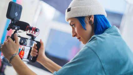 Focused Young Caucasian Female Cinematographer With Blue Hair Adjusts Camera Settings On Film Set,...