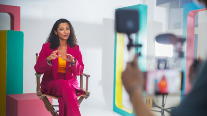 Vibrant Scene Captures A Black Female Host In A Pink Suit, Engaging Animatedly In A Colorful Studio...