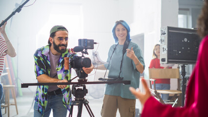 Young Caucasian Male And Female Filmmakers Collaborate On A Film Set, Adjusting Camera Equipment With Enthusiasm, Surrounded By Diverse Crew Members In A Bright, Modern Studio Environment. - 792604835