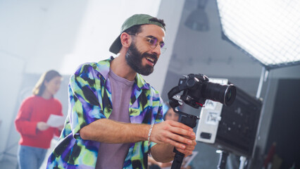 Young Male Cinematographer With Beard, Wearing A Colorful Jacket And Cap, Operates A Camera In A...