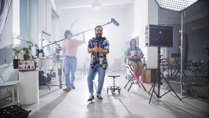 Young Diverse Team Of Filmmakers In A Bright Studio, With A Male Caucasian Videographer Holding A Camera, Surrounded By Lighting Equipment And Colleagues Reviewing Production Notes.