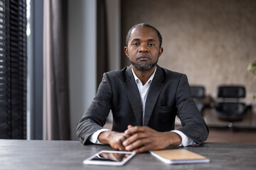 A confident African American businessman sits at a desk in a well-lit modern office, looking...
