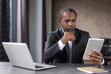 An African American businessman in a suit working intently with a tablet and laptop at his office...