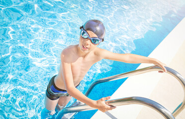 Child (boy) in cap and sport goggles on ladder handrails (stairs) in to the swimming pool, smiling...
