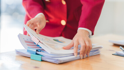 A woman is standing at a desk with a pile of papers in front of her. She is reaching for a piece of...