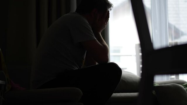 A man in despair rocks on a sofa with his face in his hands, in a dark room with light from a window and an empty alcohol bottle nearby.