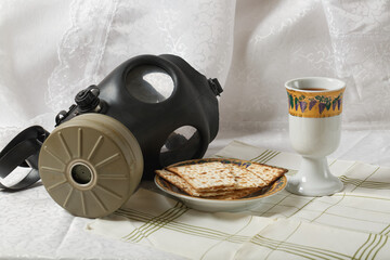 Gas mask, matzah on a plate and an ornate cup on a table with a striped cloth. The backdrop is a white lace curtain. The gasmask contrasts with the food and drink. Israeli Gas Mask. Radioactive attack