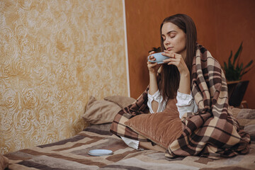 Dreamy woman wrapped in warm plaid enjoying hot cup of tea on bed in cozy room