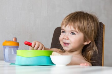 Adorable toddler girl sitting in high chair and joyfully eating healthy breakfast