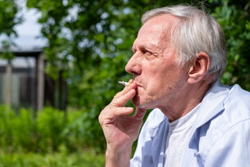 Elderly man smoking outdoors, with a contemplative look, surrounded by green foliage in a peaceful setting.