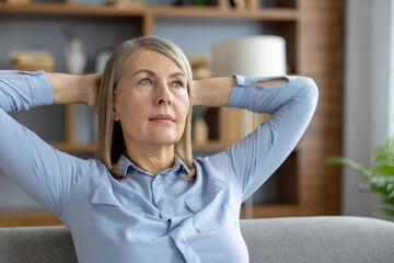 Mature woman in a blue shirt relaxing on a sofa with hands behind her head, looking thoughtful and...