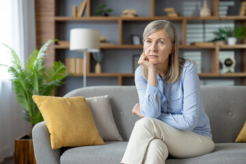 A thoughtful looking mature woman sitting in her cozy living room, gazing into the distance. The...