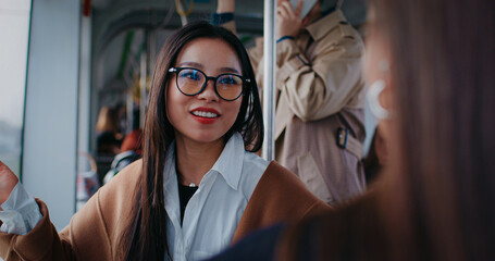 Two Asian women talking with each other in public transport. Girl with glasses listening carefully...