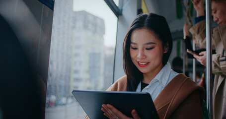 Pretty Asian woman sitting next to window in public transport. Using tablet device with big screen. Looking around afraid not to miss stop. Smiling with joy after something positive happened on gadget