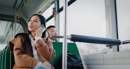 View from below of Asian woman with backpack talking actively on mobile device. Concept of remote communication. Looking around to observe urban beauty. Public transport. People in background.