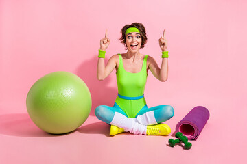 Full size portrait of astonished sporty girl sit floor indicate fingers up empty space promo isolated on pink color background