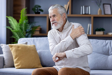 Senior gray-haired man sitting on sofa at home and holding arm above shoulder, suffering from pain...