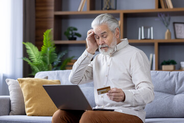 Worried senior gray-haired man sitting on sofa at home with laptop, holding head, having credit...