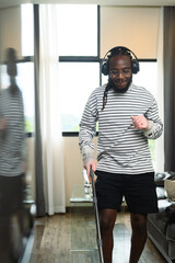 Portrait of happy African man in headphone dancing and singing while cleaning living room