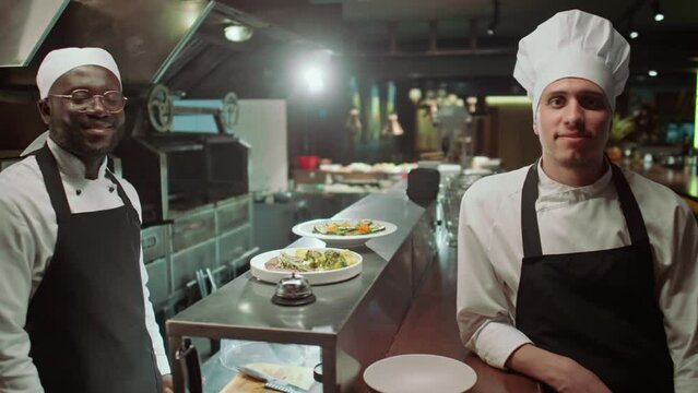 Portrait of young Caucasian and Black male chefs wearing aprons standing by food served on dishes in restaurant kitchen, smiling and posing together for cameraPortrait of young Caucasian and Black mal