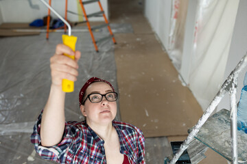 A woman in glasses and a plaid shirt is concentrating on painting the ceiling with a roller in a bright room.