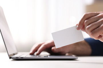 Man with laptop holding blank business card at white table indoors, closeup
