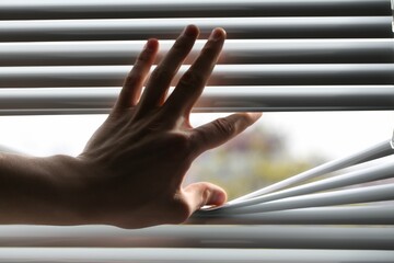 Man separating slats of white blinds indoors, closeup