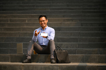 Cheerful businessman sitting on stairs of office building and showing smartphone with blank screen - 792677205