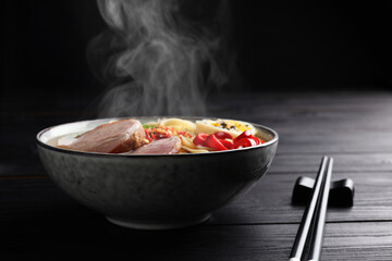 Delicious hot ramen in bowl and chopsticks on black wooden table, closeup. Noodle soup