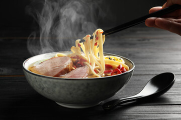 Woman eating hot ramen with chopsticks at black wooden table, closeup. Noodle soup