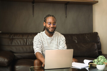 Pleased African man sitting on couch working with laptop or chatting in social