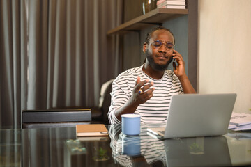 Young African man having serious phone conversation and working with laptop at home