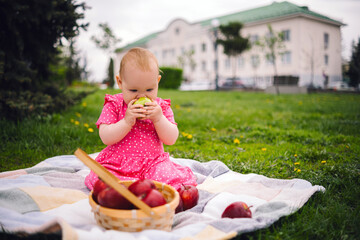A toddler in a pink dress sits on a blanket in the grass, taking a bite from a fresh apple.