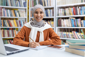 A cheerful Muslim female in a hijab is seen studying in a library, surrounded by books, with a laptop and smartphone on her desk, showcasing the concept of multicultural education.