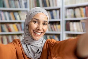 A cheerful young woman in a hijab takes a selfie with a smartphone, standing before rows of books...