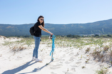 A conscious female helps clean the beach by picking up a blue piece of trash on a sunny day,...