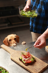 A man prepares avocado and ham bruschettas, with a loyal pet watching, creating a cozy home cooking scene.
