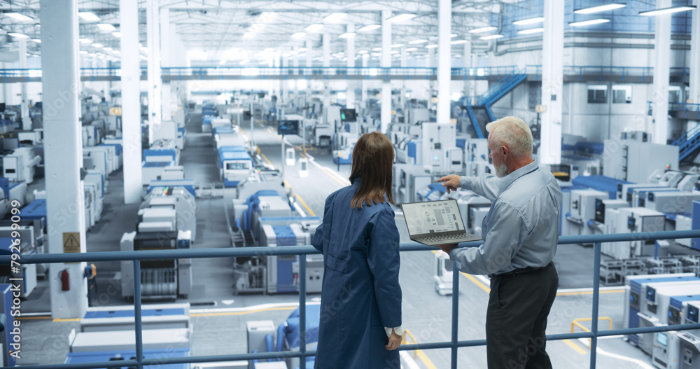 Poster Two Multiethnic Engineers Standing on a Platform, Using Laptop Computer at an Electronics Factory. Machines are Undergoing Maintenance, Specialists Monitoring the Progress Through Online Software