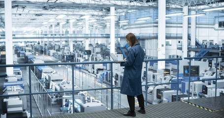 Experienced Female Engineer Standing on a Platform, Using Laptop Computer and Overlooking Production at a Modern Automated Electronics Manufacture with AI Support. Factory Manager at Work